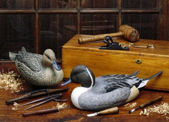 Two detailed wooden duck carvings sit on a wooden table, surrounded by carving tools and wood shavings. A wooden toolbox, mallet, and plane are in the background against a paneled window.