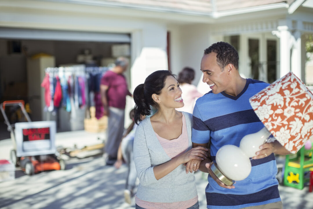 A smiling couple holds a lamp and walks at a garage sale in a driveway. Behind them, a variety of items are displayed, with a "Yard Sale" sign visible and people browsing in the background.