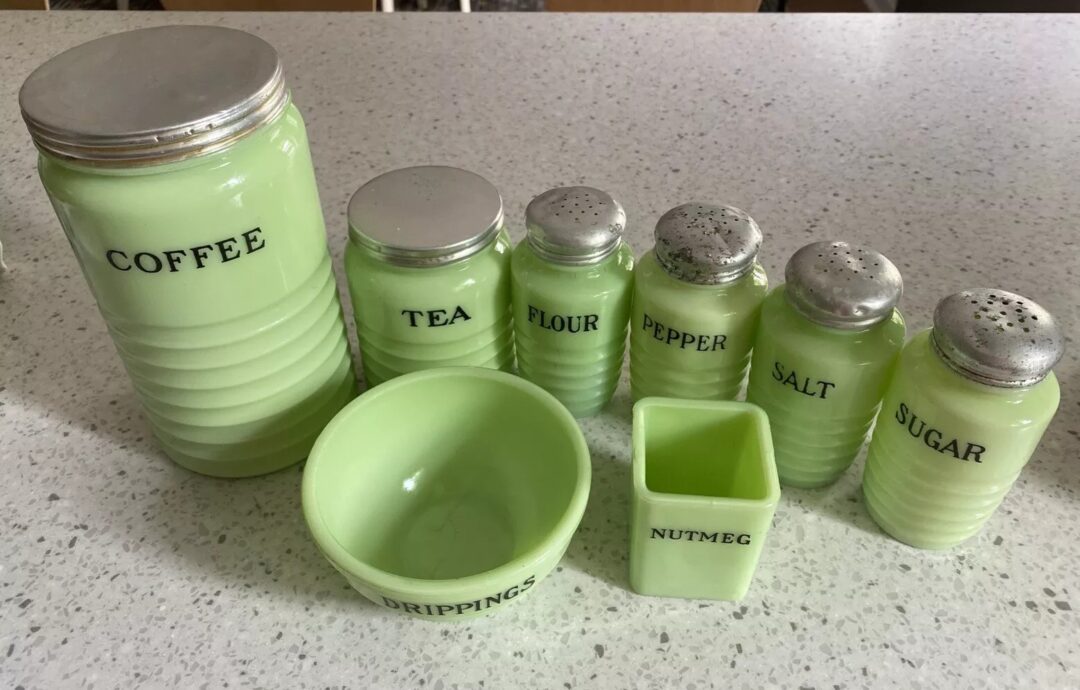 A set of vintage green glass kitchen containers with metal lids labeled for coffee, tea, flour, pepper, salt, sugar, nutmeg, and drippings. They are arranged on a speckled countertop.