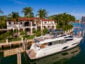 A large white yacht is docked at a waterfront property featuring a luxurious mansion with red-tiled roofs and surrounded by palm trees. The sky is clear, and the waters are calm, reflecting the greenery and buildings.