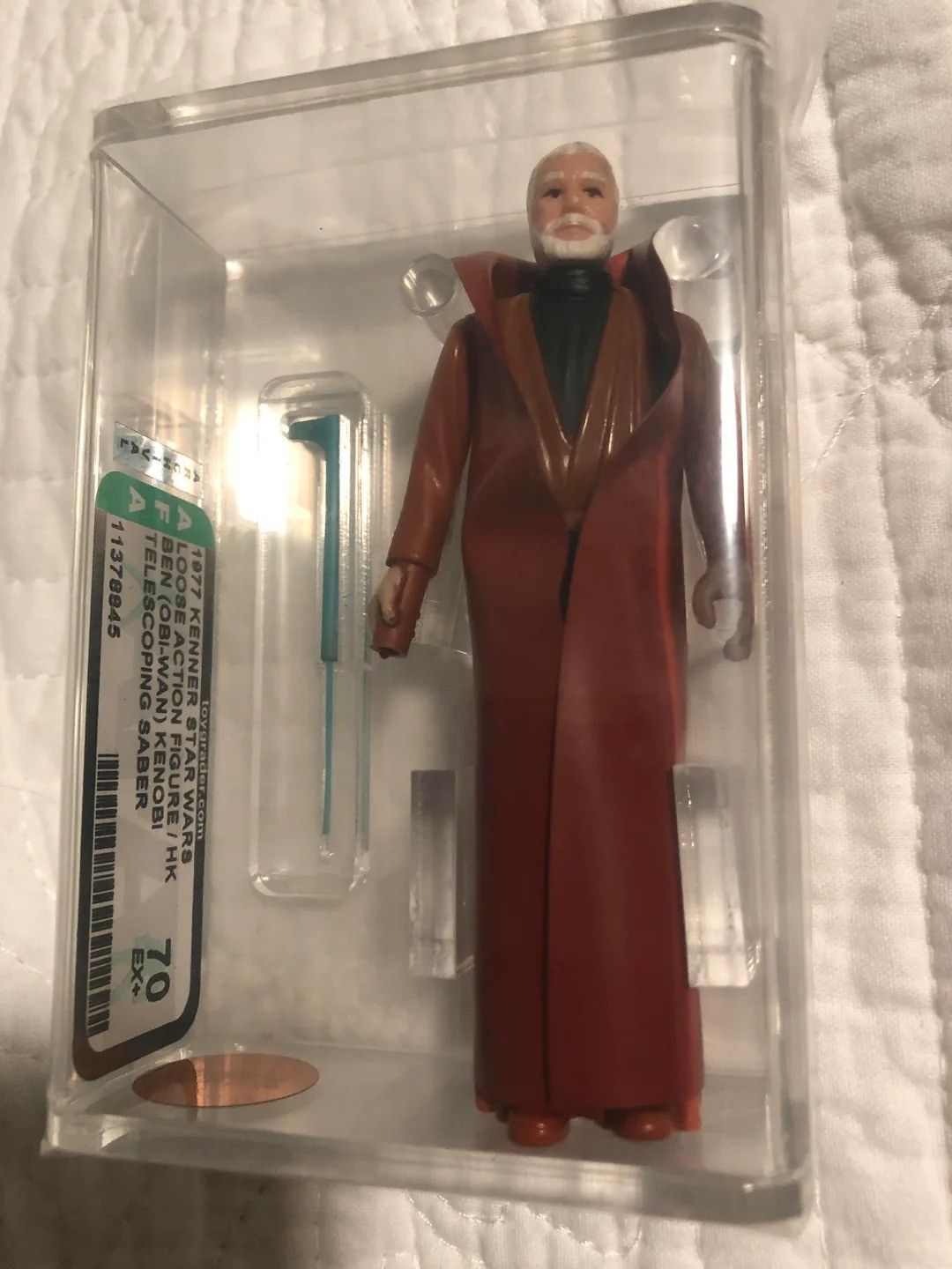 A vintage Star Wars action figure in a clear acrylic case, featuring a bearded character in a brown robe. A blue lightsaber accessory is included beside the figure. The background is a white textured surface.