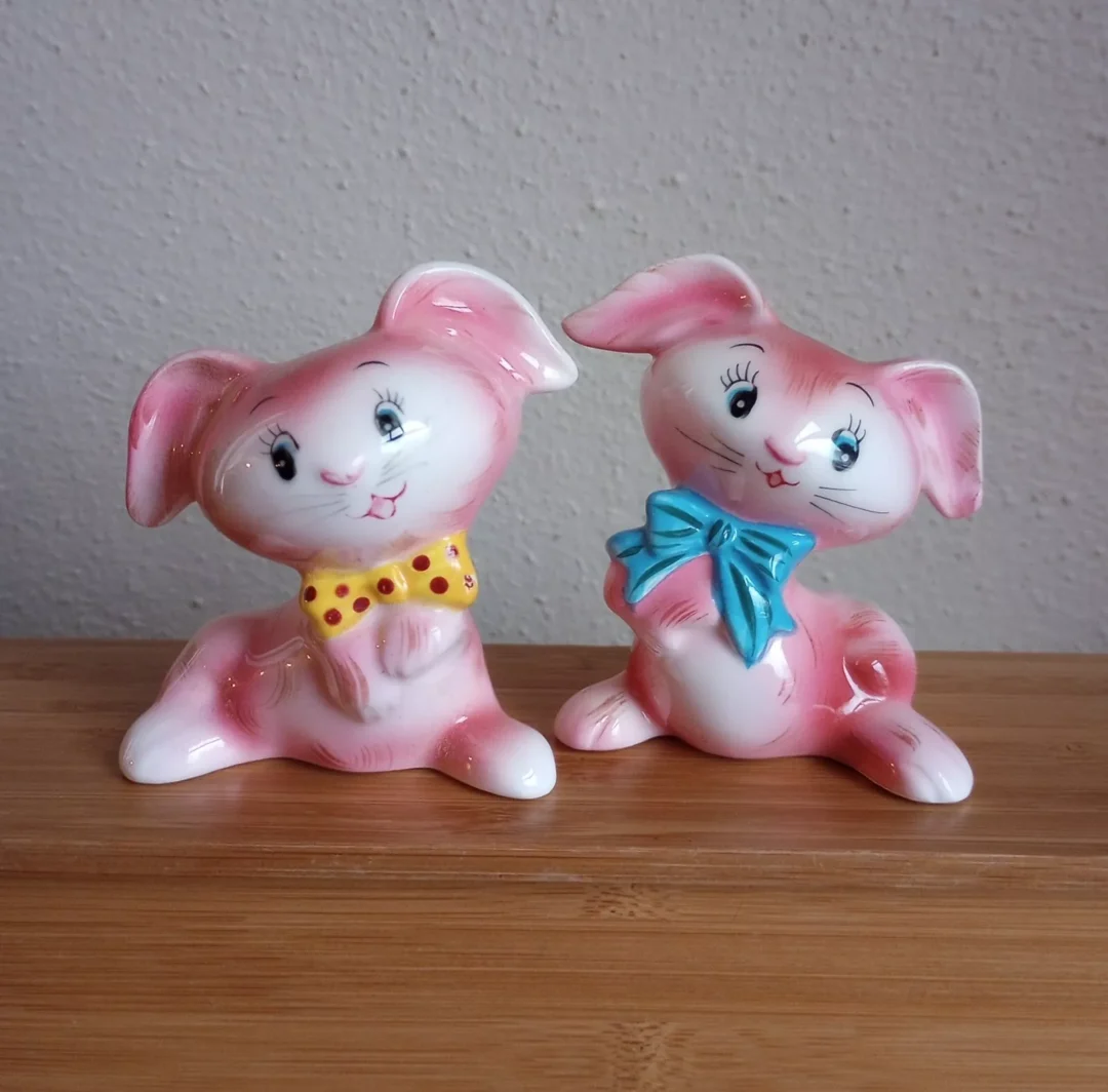 Two pink ceramic bunny figurines with playful expressions. One has a yellow polka-dotted bow tie, and the other has a blue bow. They're sitting on a wooden surface against a light-colored wall.