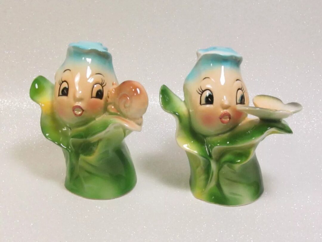 Two figurines with blue hats and rosy cheeks, each holding a leaf. The left one has a snail on its hand, and the right one holds a small flower. The figures have wide eyes and are set against a sparkling white background.