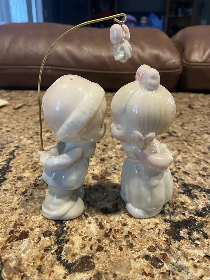 Two ceramic figurines of a boy and girl leaning in for a kiss. The girl wears a pink bow, and a wire with a tiny butterfly connects them. They stand on a speckled countertop with a brown couch in the background.