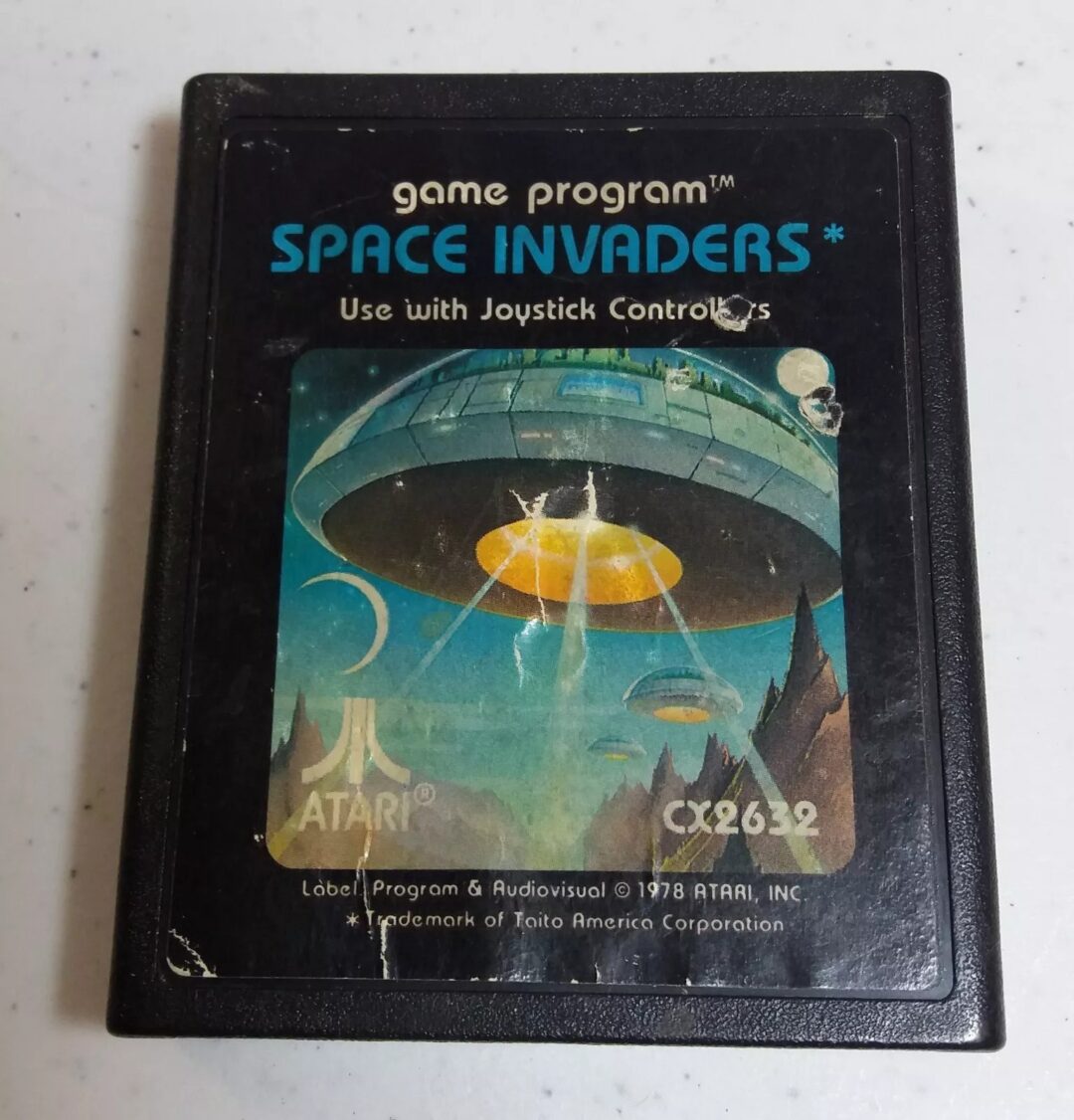 Close-up of an Atari game cartridge for "Space Invaders" with artwork of a UFO beaming light onto a landscape. Text includes "game program™ SPACE INVADERS*" and "Use with Joystick Controllers." Atari and CX2632 are noted.