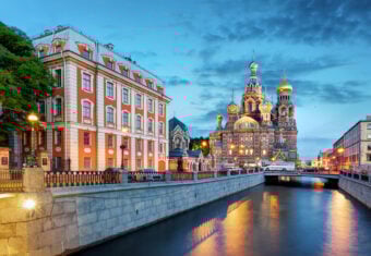 View of the colorful Church of the Savior on Spilled Blood and a nearby historic building along a canal in St. Petersburg, Russia, at dusk. The sky is blue and partially cloudy, with streetlights casting a warm glow on the scene.