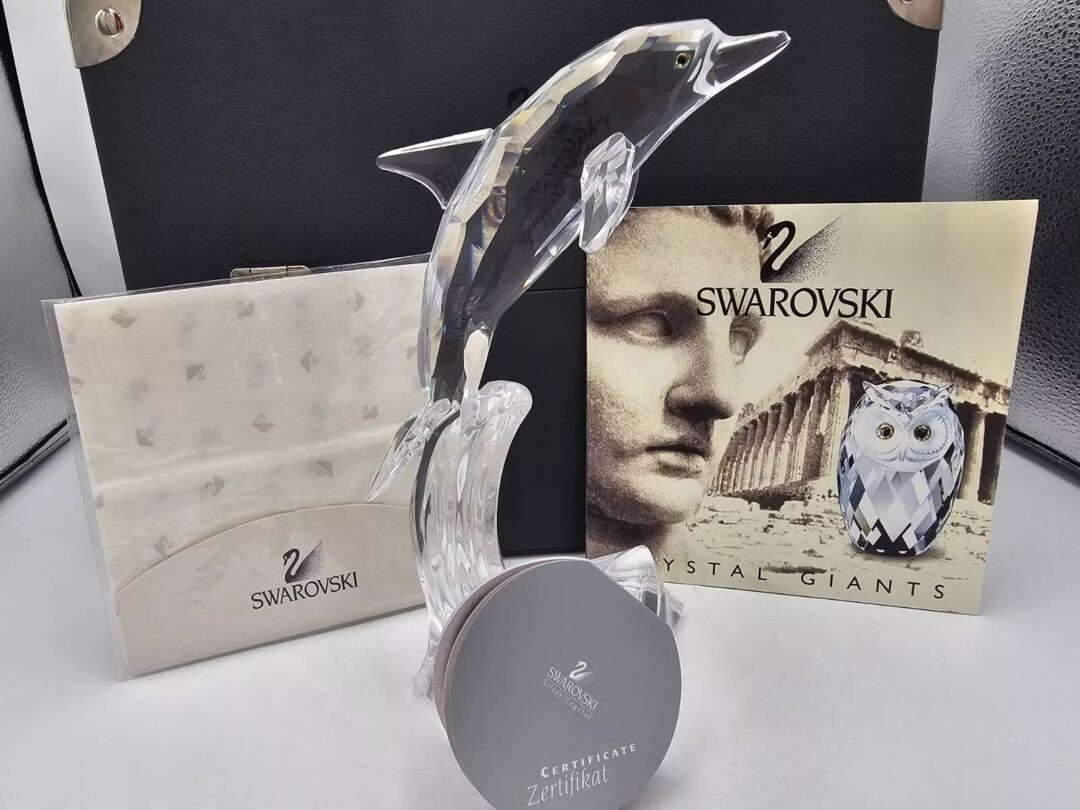 A crystal dolphin sculpture leaps from a base with a "Zertifikat" certificate. Behind it are Swarovski packaging and a booklet featuring a mosaic pattern and an owl design.