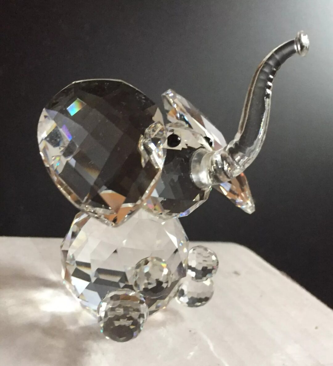 A small, intricately crafted glass elephant figurine with a raised trunk sits on a flat surface. The figure reflects light, showcasing its faceted surface, creating a sparkling effect against a dark background.