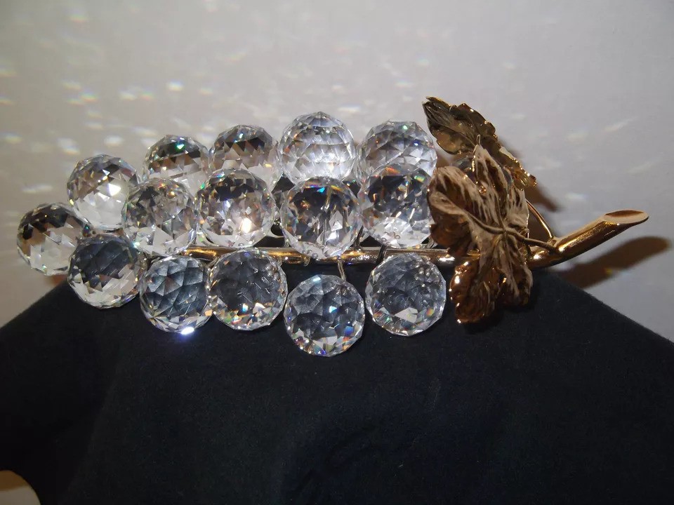 A decorative grape cluster made of clear crystal spheres is attached to a gold-tone stem with detailed gold-tone leaves, displayed against a dark background.