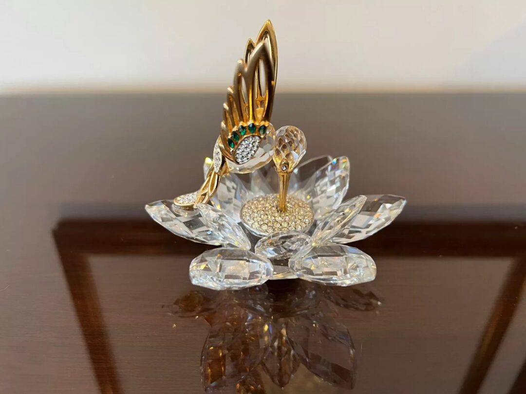 A decorative crystal swan rests on a lotus-shaped base. The swan features a gilded neck, a metallic wing with an emerald-green accent, and sparkling embellishments, set on a glossy wooden surface.