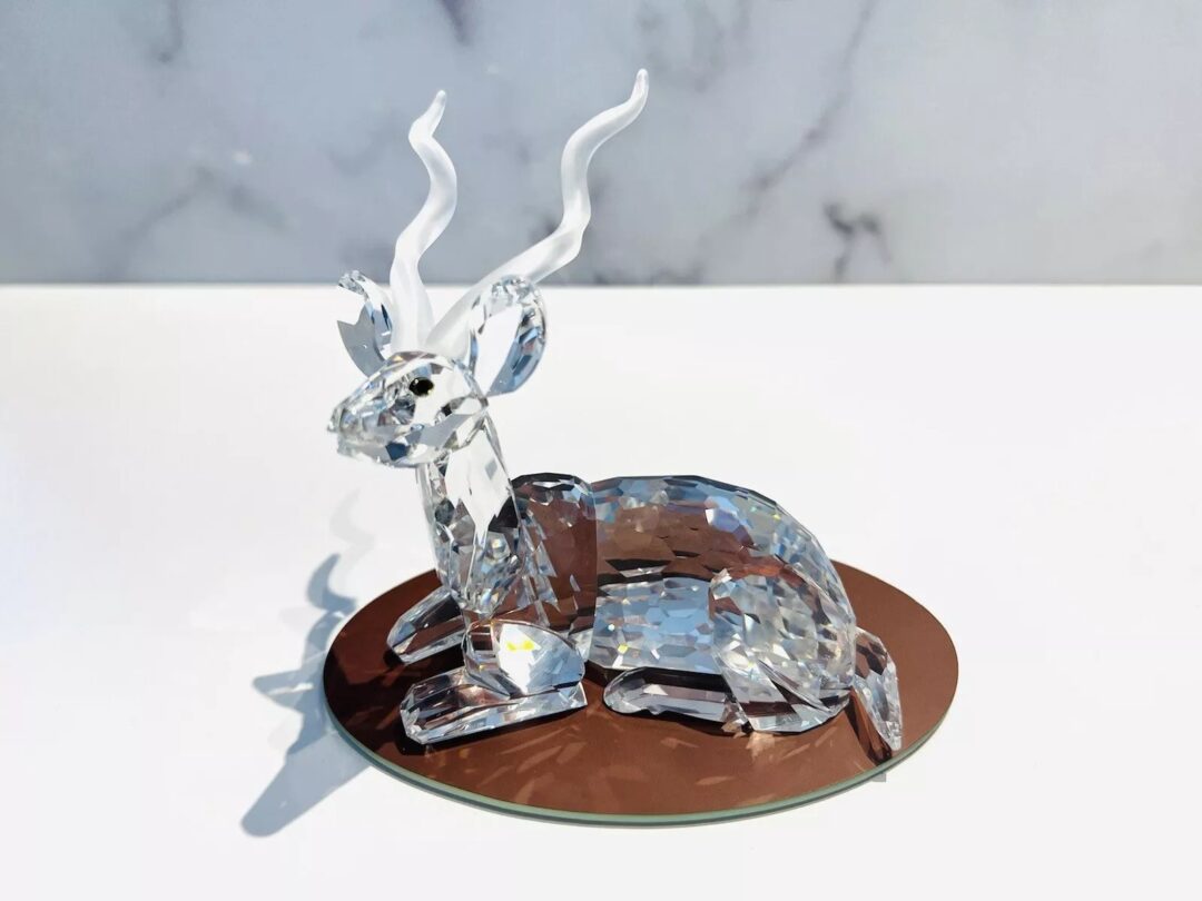 A crystal sculpture of a deer with long antlers, sitting on a round brown base. The transparent material reflects light, creating a sparkling effect. The background is a grayish marble pattern.