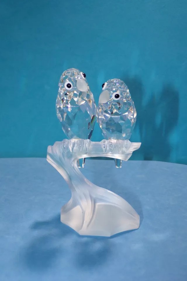 Two crystal bird figurines perched on a transparent stand against a blue background. The birds have faceted designs, reflecting light, and each has distinct, black eyes. The stand has a wave-like shape, adding an elegant touch.