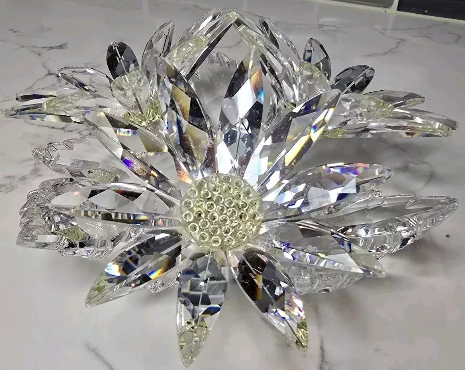 A decorative crystal flower with layered, faceted petals and a textured center, placed on a marble surface. The petals catch and reflect light, creating a sparkling effect.