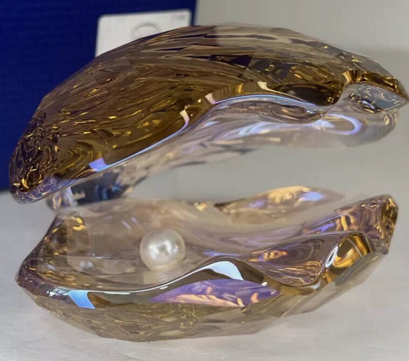 A decorative glass oyster displaying a white pearl inside. The glass oyster features a translucent, amber-hued shell with intricate details and a polished finish, set against a minimalist background.