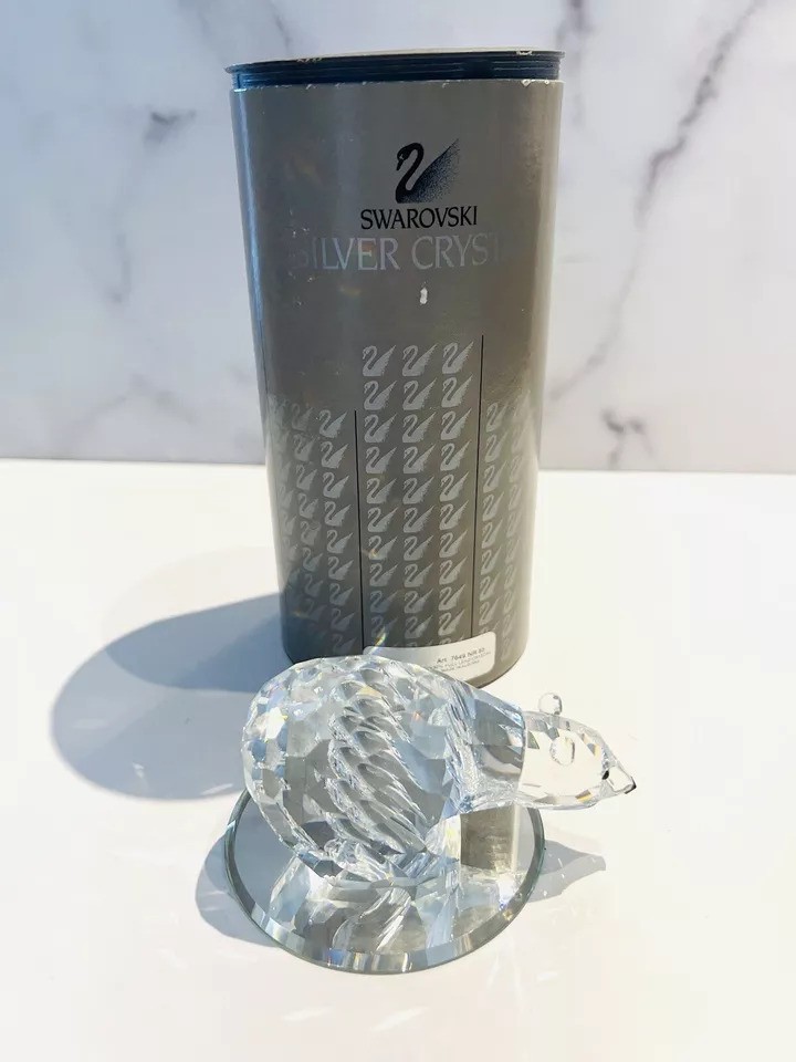 A Swarovski crystal figurine of a hedgehog stands in front of a tall, cylindrical Swarovski Silver Crystal box. The figurine is intricately detailed and sits on a reflective surface.