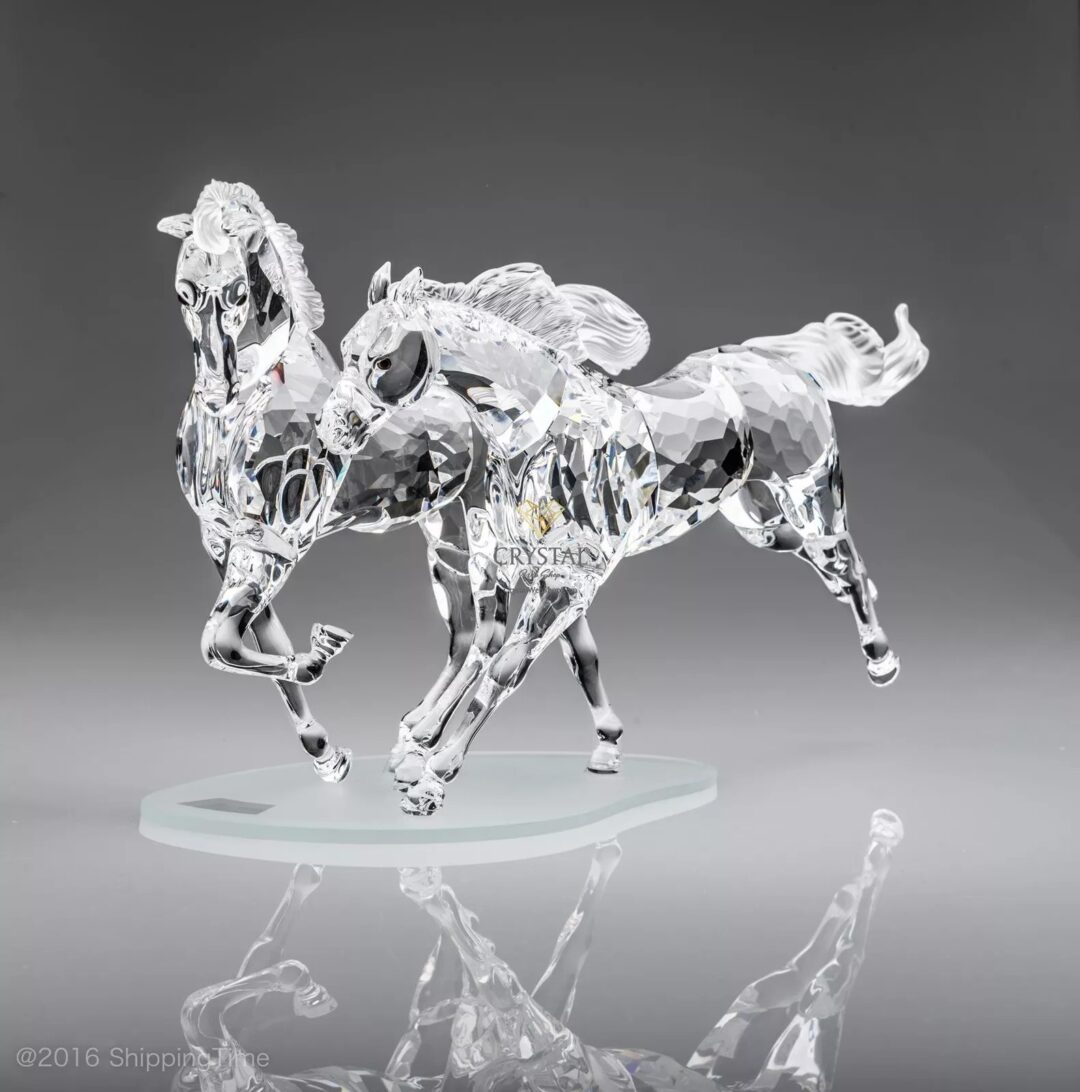 A crystal figurine depicting two galloping horses with intricate detailing and a smooth, reflective finish, set against a gray background.