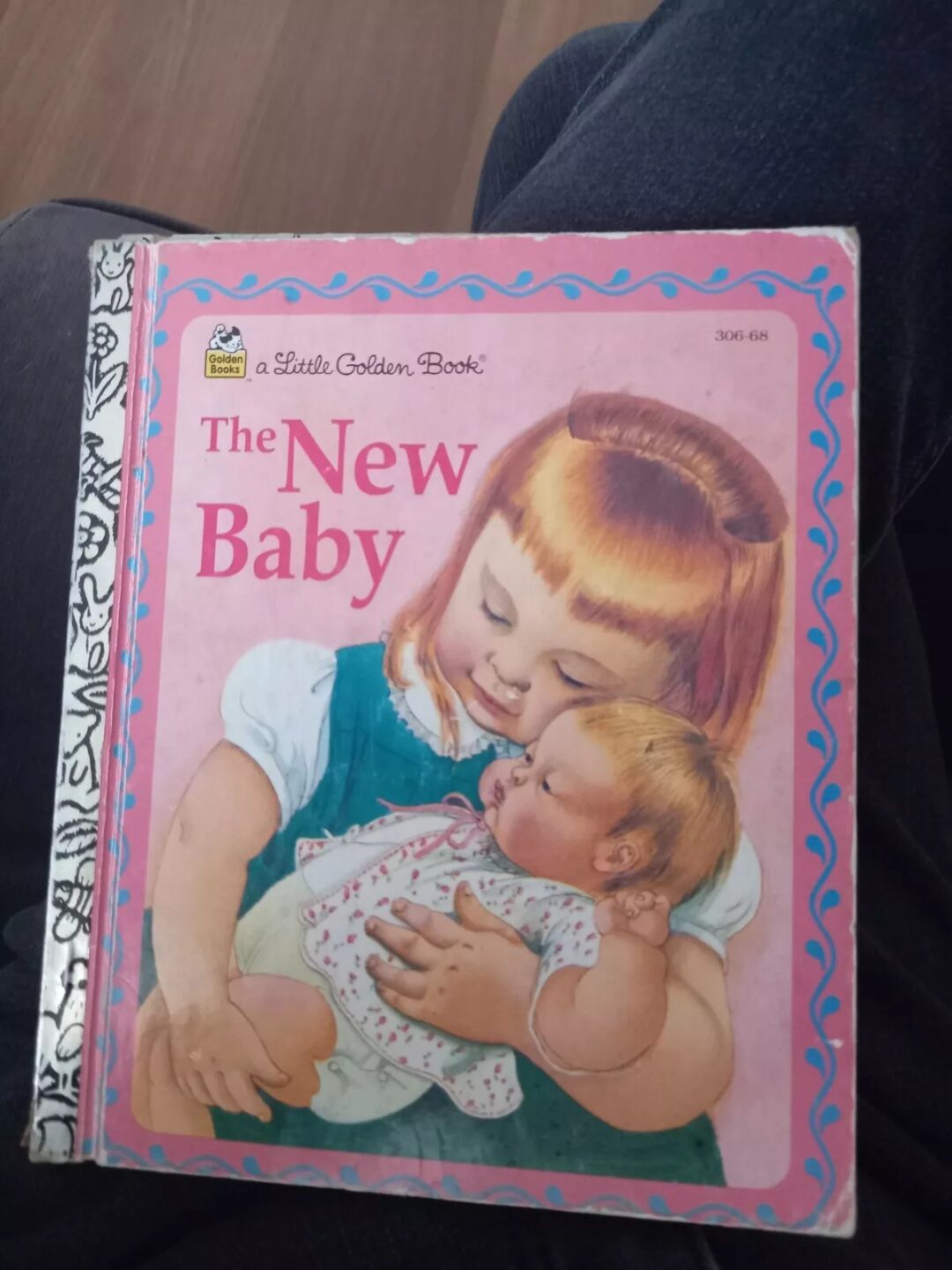 A Little Golden Book titled "The New Baby" with a pink cover. It features an illustration of a young girl holding a baby, both with light-colored hair. The book's spine has a black and white decorative pattern.