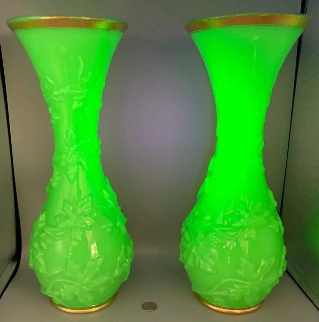 Two tall, glowing green glass vases with floral patterns are displayed side by side. Each vase has a slightly flared top and gold trim. A small coin at the base provides scale.