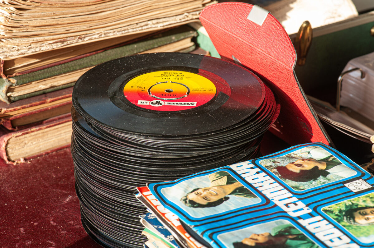 A stack of vintage vinyl records sits on a table next to old books and magazines. A colorful magazine with images of people is partially visible in the foreground. The setting has a nostalgic, retro vibe.