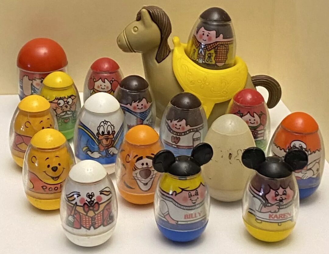 A group of colorful egg-shaped toys with cartoon faces, featuring characters wearing various outfits. A toy camel stands behind them. The background is a plain beige surface.