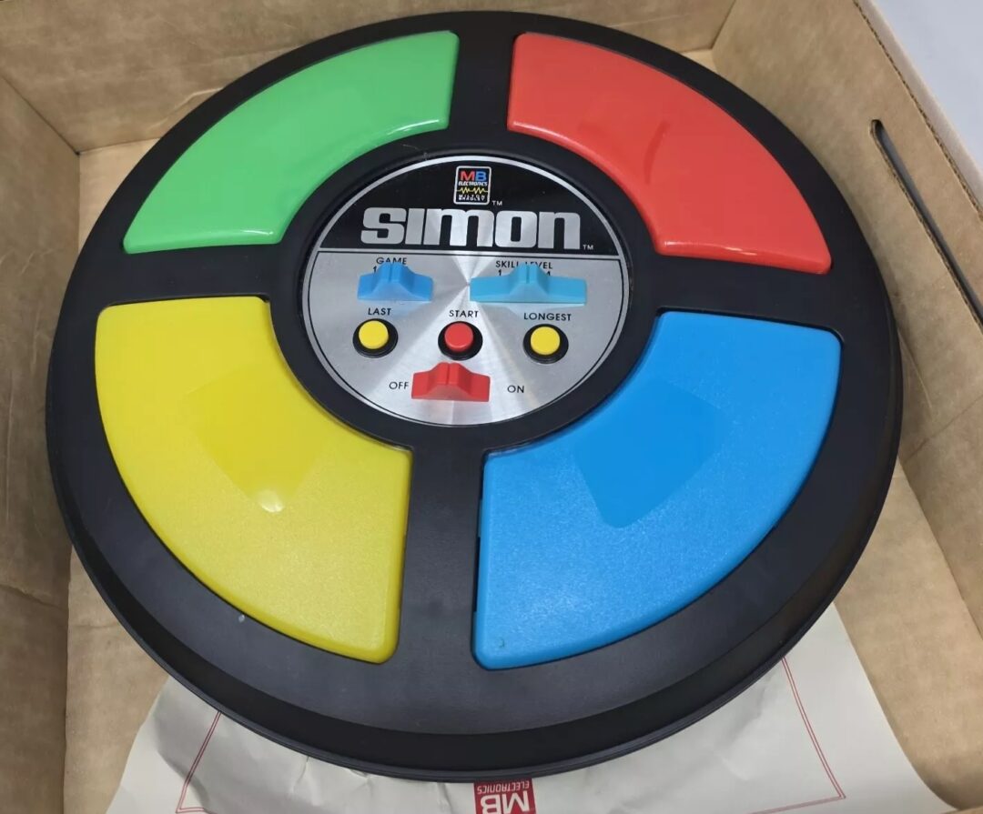 A vintage Simon electronic memory game in a cardboard box. The game features four large colored buttons—green, red, blue, and yellow—and a central control panel with switches. The MB logo is visible on the box.