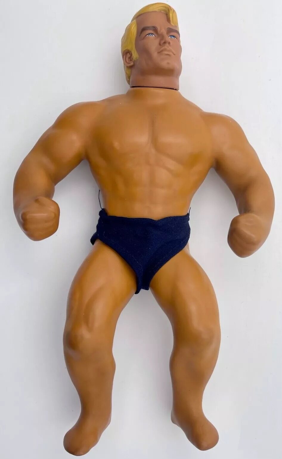 A vintage action figure with muscular build, wearing dark blue shorts. The figure has blonde hair and slightly bent arms, positioned against a plain white background.