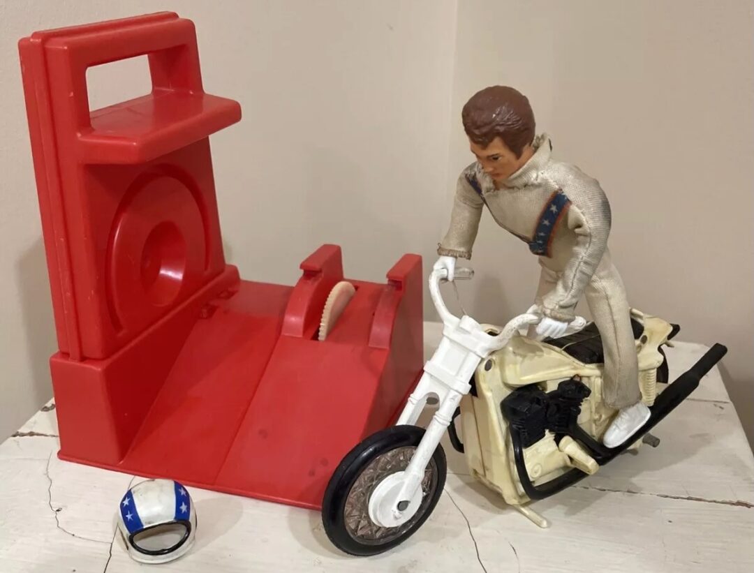 A toy motorcycle with a rider figure in a white outfit is on a white table. Next to it is a red ramp launcher and a small blue and white helmet. The background is a plain wall.