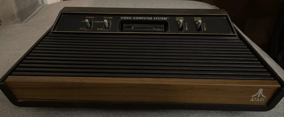 A vintage Atari video game console with a woodgrain panel is displayed on a gray surface. The console features switches for power, TV type, and game selection.