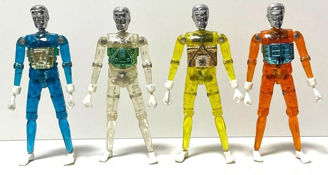 Four colorful, transparent robot action figures stand side by side. Each has a different color scheme: blue, clear, yellow, and orange. Their bodies reveal visible internal circuit designs, and all have silver heads and white hands.