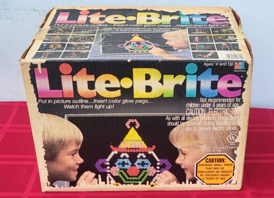 A vintage Lite-Brite toy box featuring colorful graphics of two children facing each other with a completed design of a smiling clown. The box highlights the toy's function of creating illuminated pictures using colored pegs.