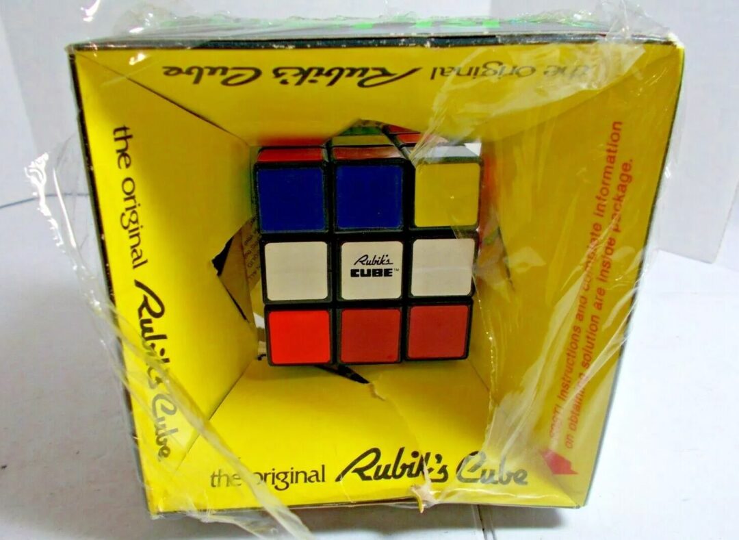 A Rubik's Cube encased in a damaged yellow box. The packaging is partially torn and wrapped with clear plastic. The Rubik's Cube is partially solved, showcasing various colored sides like red, blue, and white.