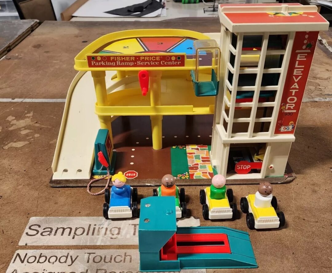 A vintage Fisher-Price Toys playset depicting a multi-level parking ramp with an elevator. Four small toy figures are seated in colorful cars. The playset is on a table alongside a Fisher-Price logo sign.