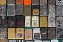 A collection of various Zippo lighters is displayed. Each lighter features unique designs, including logos, symbols, and artwork in different colors and finishes, arranged in a grid.