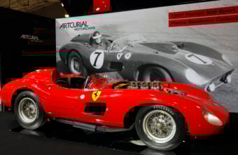A classic red Ferrari 335 Sport car is on display in front of a large black and white photo of the same model racing. The car features open wheels and a sleek, vintage design. The display is labeled "Artcurial Motorcars" and "Pierre Bardinon Collection.