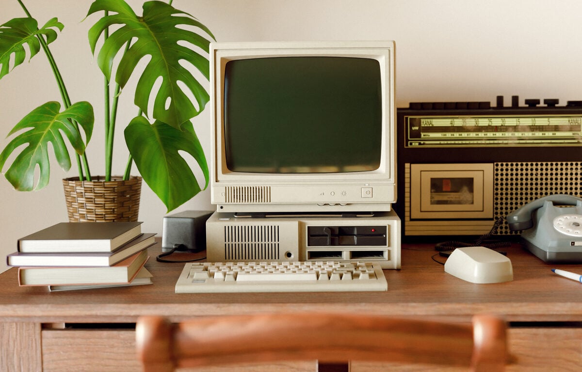 A vintage desk setup featuring an old computer with a CRT monitor, floppy disk drives, a rotary dial telephone, a tabletop radio, a stack of books, a potted monstera plant, and a computer mouse on a wooden desk.
