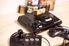 A Sega Genesis video game console with a cartridge inserted, accompanied by a joystick arcade controller and a standard controller, all placed on a light wooden surface. Game cartridges are visible in the background.