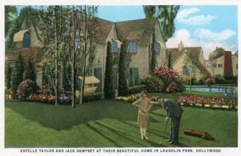 A man and woman are playing croquet on a lush green lawn in front of a large, elegant house. Colorful flowers and tall trees adorn the garden. Text at the bottom mentions they are at their home in Laughlin Park, Hollywood.