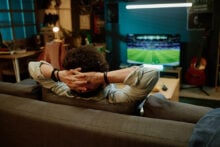 A person with curly hair is sitting on a sofa with hands behind their head, watching a soccer game on a large TV in a dimly lit room. A guitar is leaning against the wall, and a computer setup is visible in the background.