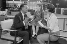 A man and woman smile at a young girl sitting between them in a department store. The man holds a wallet, and the girl laughs. The scene appears to be from the 1950s or 1960s, depicted in black and white.