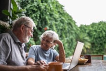Elderly couple sitting at an outdoor table covered with a tablecloth, smiling at each other. A laptop is open in front of them, with a plant and greenery in the background. A coffee mug and bowl are on the table.