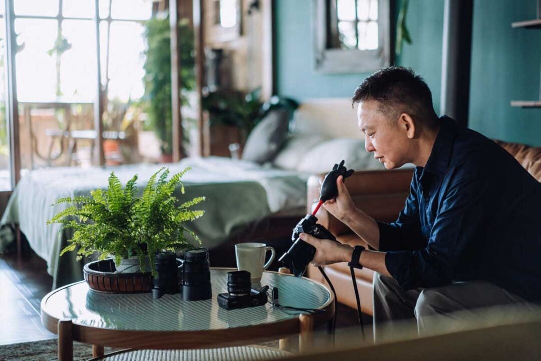 A man sits on a sofa in a cozy, sunlit room, cleaning a camera. Camera lenses, a cup, and a potted fern are on the table in front of him. A bed and large window are in the background.