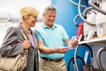 An older couple stands in an appliance store, smiling and reading a brochure while looking at a selection of vacuum cleaners displayed on a shelf.