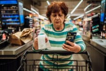 An older woman in a striped shirt stands at a grocery store checkout, holding a receipt and looking at her smartphone. She has a shopping cart in front of her, and shelves with products in the background.