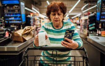 An older woman in a striped shirt stands at a grocery store checkout, holding a receipt and looking at her smartphone. She has a shopping cart in front of her, and shelves with products in the background.
