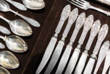 A set of ornate silverware featuring intricate patterns on the handles. The image shows seven knives and six spoons, all aligned neatly on a dark background. A single fork rests above the knives.