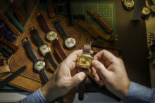 Hands holding a vintage square watch with a brown leather strap over a table displaying various watches and straps. The table has a leather mat and a green cutting board, and the watches have different strap colors and designs.