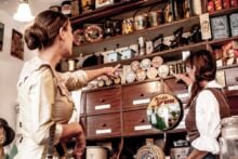 Two women in vintage clothing point at tins and containers on shelves in an old-fashioned store, surrounded by wooden cabinets and retro products. The store has a nostalgic, historical atmosphere.