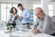 An older man with glasses works at a desk, writing on documents. Two colleagues stand in the background, discussing and looking at a tablet. The office is bright with natural light and modern decor.