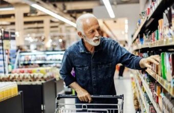 An elderly man with white hair and a beard, wearing a denim shirt, pushes a shopping cart and selects items from a grocery store shelf.