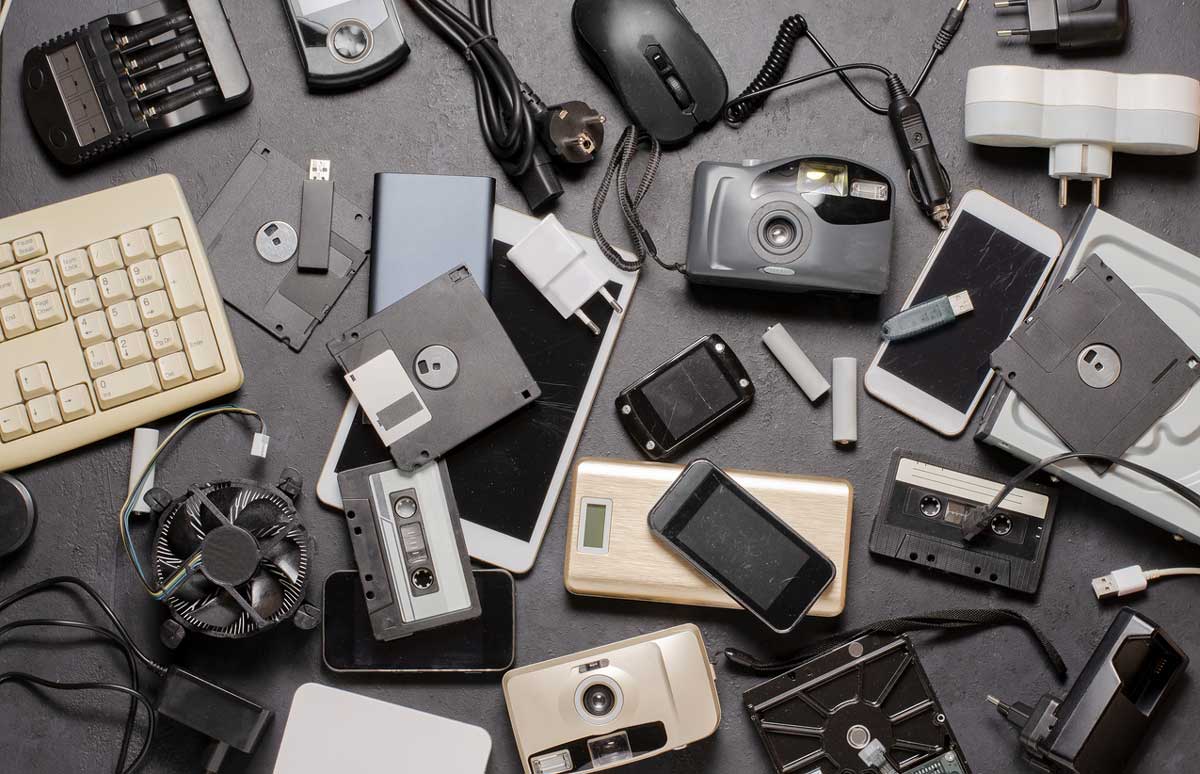 A variety of old electronic devices and accessories, including floppy disks, cassette tapes, cameras, smartphones, a keyboard, a mouse, chargers, and other miscellaneous tech items spread out on a dark surface.